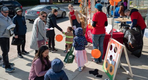 A lively community event featuring families and children. Participants engage in activities, with children playing near a game booth decorated with colorful balloons. Attendees are dressed in casual clothing, enjoying a sunny day outdoors.