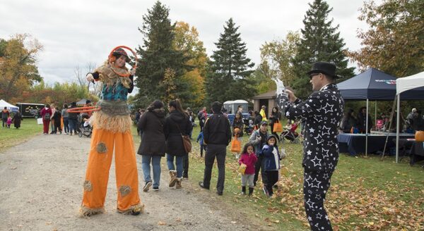 A colorful festival scene shows a stilt walker in a scarecrow costume interacting with a person in a black and white patterned outfit. Families and children enjoy the event outdoors, surrounded by trees with autumn foliage.