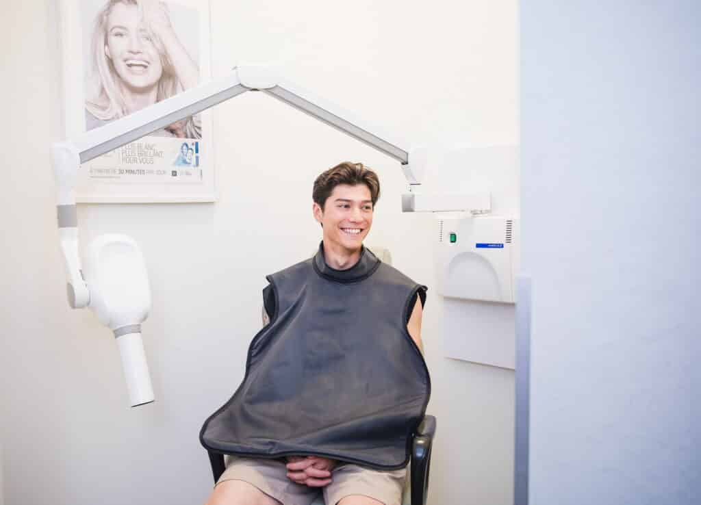 A young man is sitting in a dental chair, wearing a protective bib and smiling. A dental light is positioned above him, and a poster of a smiling woman is visible on the wall behind. The atmosphere appears friendly and welcoming, featuring the 123Dentist Smiley in the context.