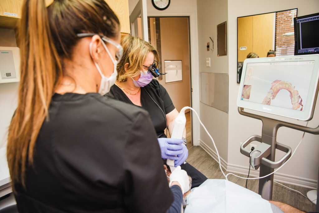 Two dental professionals are working together in a clinic, wearing masks and gloves. One is using a dental instrument, while the other observes and assists. A digital display with an image of dental scans is positioned nearby, contributing to the treatment process.