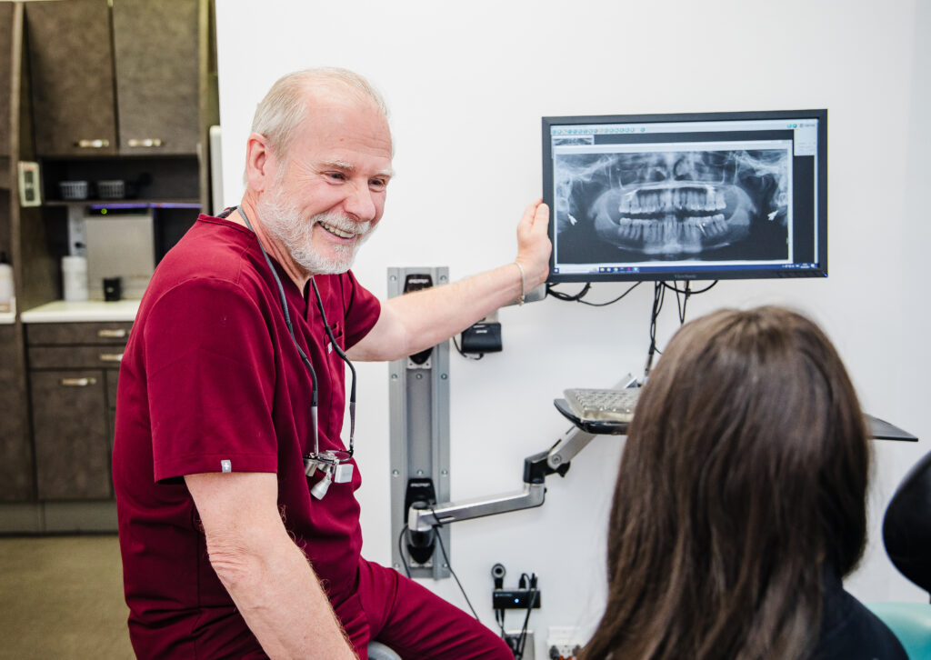 A friendly dentist in red scrubs smiles while showing a patient an X-ray on a computer monitor in a dental office. The warm atmosphere promotes trust and communication between the dentist and the patient.