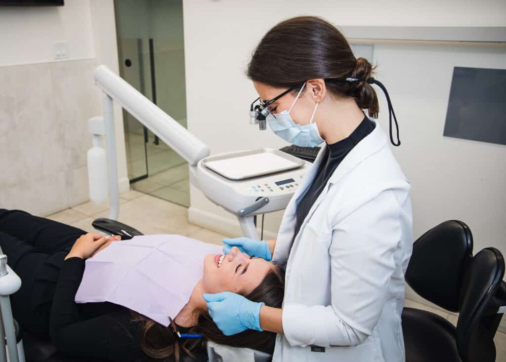 A dental professional in a white coat and mask attends to a patient in a dental chair, adjusting a bib. The office is modern, equipped with dental tools. The patient appears relaxed, ensuring a calm atmosphere during the appointment.