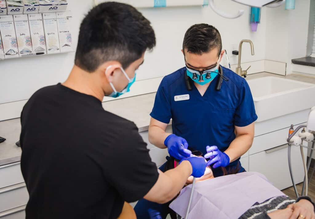 A dental professional in blue scrubs and gloves examines a patient receiving treatment in a dental office. Another individual, likely a dental assistant, observes the procedure. The scene emphasizes a clean, clinical environment focused on dental care.