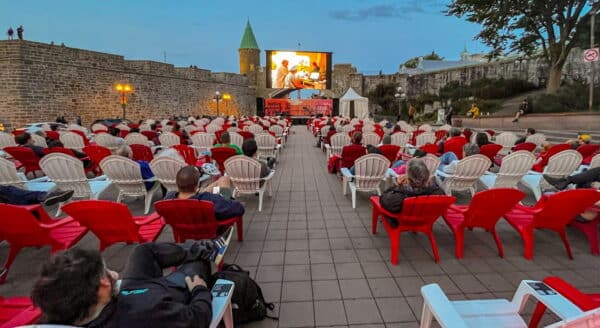 An outdoor movie screening is set up with numerous red and white chairs arranged in front of a large screen displaying a film. The scene is soft-lit during dusk, with a historic wall and trees in the background, creating a cozy atmosphere for viewers.