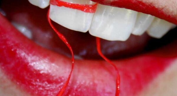 A close-up of a mouth with bright red lipstick, showing white teeth and strands of red thread resembling blood dripping from the corners.