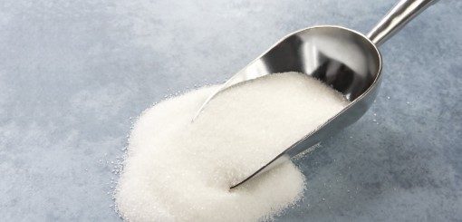 A metal scoop is shown partially filled with granulated white sugar, with a small pile of sugar scattered underneath it on a smooth surface.