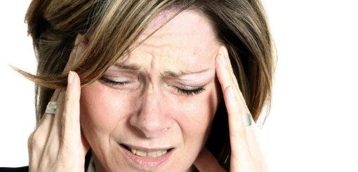 Une femme aux cheveux mi-longs se tient la tête à deux mains, semblant angoissée ou souffrante. L'expression de son visage suggère une gêne, probablement due à un mal de tête ou au stress. L'arrière-plan est simple et clair.