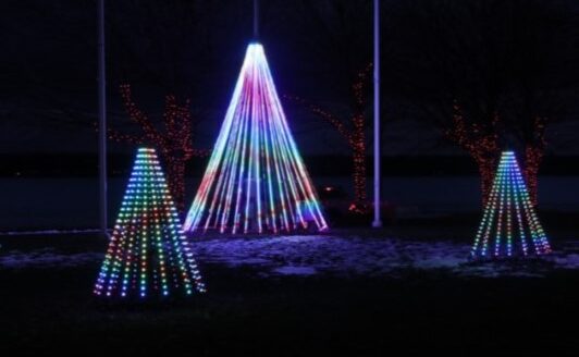 Colorful light displays shaped like Christmas trees are arranged in a dark outdoor setting, with bright neon lights creating a festive atmosphere. The background features a river and trees adorned with additional lights.