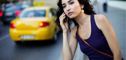 A young woman with dark, wavy hair stands on a city street, holding a smartphone to her ear, while a yellow taxi is parked nearby. She is wearing a sleeveless purple top and appears engaged in conversation.