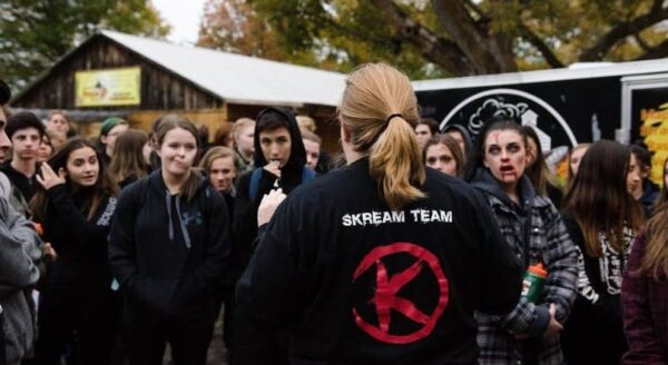A group of teenagers listens intently to a leader wearing a black "Scream Team" sweatshirt, as they gather outdoors in a fall setting with trees in the background. The atmosphere suggests a team briefing or event preparation.