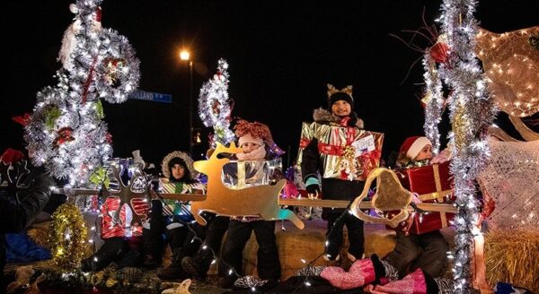 A festive parade float featuring performers dressed in colorful costumes, surrounded by holiday decorations and lights. The scene is set at night, creating a vibrant and cheerful atmosphere.