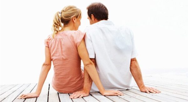 A couple sits closely together on a wooden dock, facing away from the camera, enjoying a quiet moment. The light background suggests a serene outdoor setting.