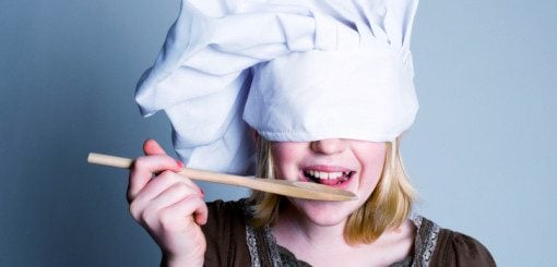 A child wearing a chef's hat playfully holds a wooden spoon while pretending to taste food. Their eyes are covered with the hat, adding a sense of fun and imagination to the scene.
