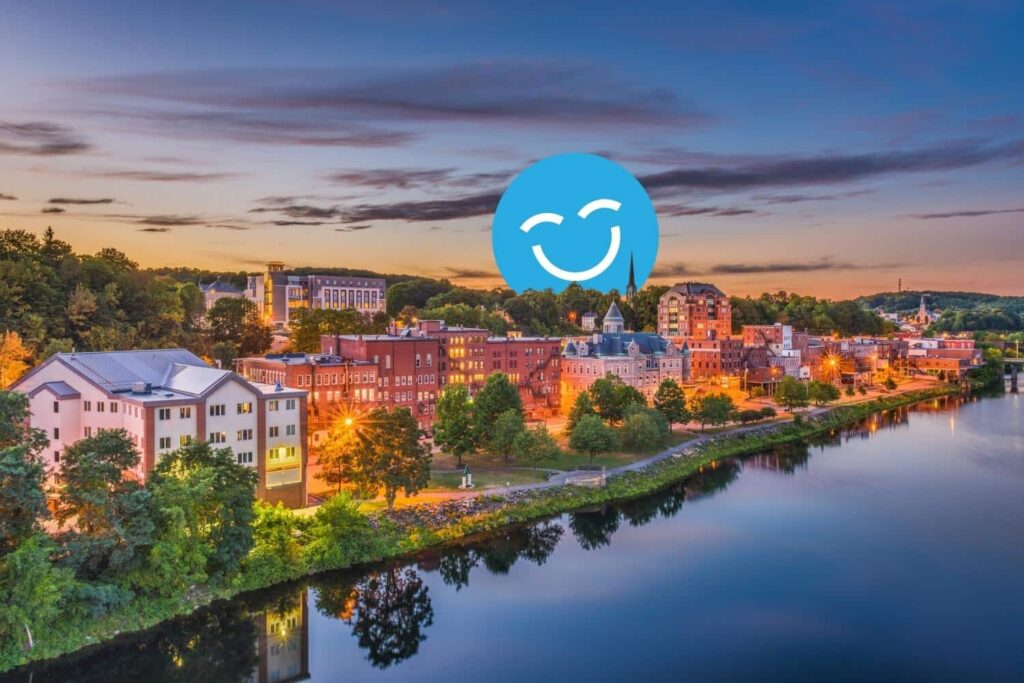 Une vue pittoresque d'une ville au bord de la rivière au coucher du soleil, avec des bâtiments charmants le long de l'eau, des arbres et des reflets. Une icône de visage souriant bleu se superpose à l'image, ajoutant un élément joyeux à la scène.
