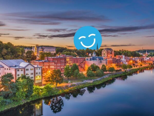 A picturesque view of a riverside town at sunset, featuring charming buildings along the water with trees and reflections. A blue smiley face icon overlays the image, adding a cheerful element to the scene.