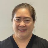 A woman with medium-length black hair smiles at the camera. She is wearing a dark-colored shirt and has small earrings. The background is light gray, suggesting a professional or clinical setting.
