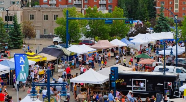 A bustling outdoor market with numerous vendor tents, colorful canopies, and a crowd of shoppers enjoying the vibrant atmosphere. Trees and buildings are visible in the background, creating a lively community setting.