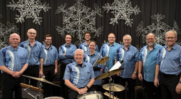 A group of twelve musicians wearing matching blue shirts pose for a photo, with one member seated at a drum set. The background features decorative snowflakes, creating a festive atmosphere.