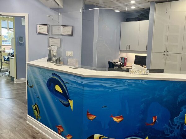 Reception area in a dental office with an ocean-themed counter featuring colorful fish murals, white cabinets, and soft blue walls.