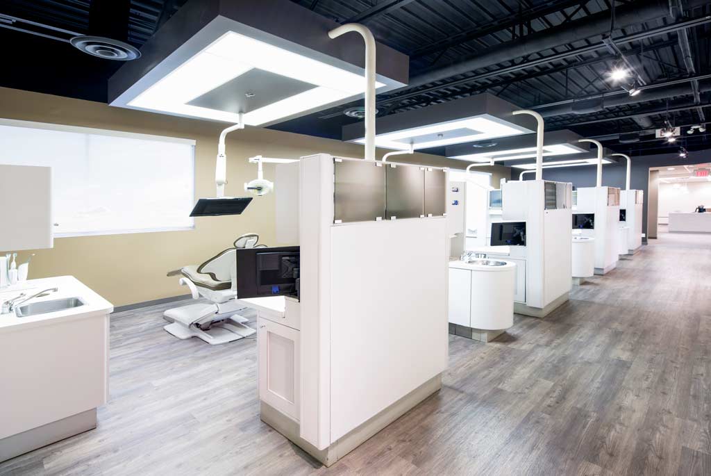 Modern dental clinic interior with several workstations, white partitions, and dental chairs. The space features sleek design, bright overhead lighting, and a clean, organized layout.