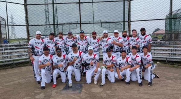 A group of 22 individuals poses together on a baseball field, wearing matching white uniforms with red and black accents. They stand in front of a chain-link fence with bleachers in the background, smiling and showcasing camaraderie as a sports team.