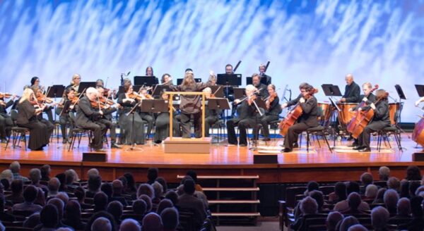An orchestra performs on stage, with musicians playing various instruments. The conductor leads the ensemble, while an audience observes from their seats. The background features a colorful, wavy backdrop, enhancing the concert atmosphere.