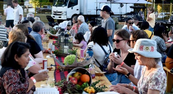 A vibrant outdoor event featuring a diverse group of people seated at long tables adorned with fruits and vegetables. Participants engage in conversation and enjoy the atmosphere as a truck and stage are set up in the background, creating a lively communal experience.