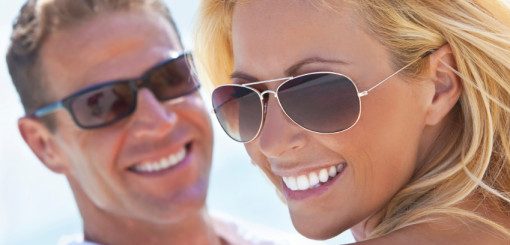 A smiling couple wearing sunglasses enjoys a sunny day, with the woman in focus and the man slightly blurred in the background, conveying a sense of happiness and relaxation.