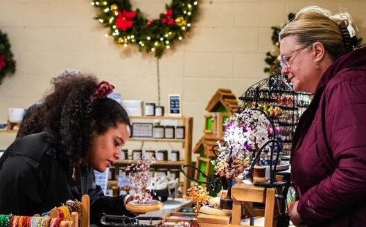 Two women are engaged in a conversation at a retail store, with one woman showing interest in various handmade items. Holiday decorations, including a wreath, are visible in the background, enhancing the festive atmosphere.