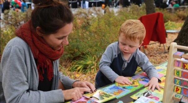A woman and a young boy are engaged in an interactive activity at a table, exploring colorful books and educational materials outdoors. The scene is lively, with people and greenery in the background.