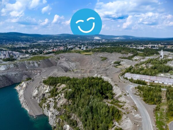 Aerial view of a large quarry surrounded by greenery, with a small lake nearby. A city skyline and hills are visible in the background. A blue smiley face emoji is overlaid on the sky.