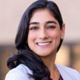 A smiling woman with long dark hair, wearing a light-colored blazer, stands in front of a neutral background, projecting confidence and warmth.