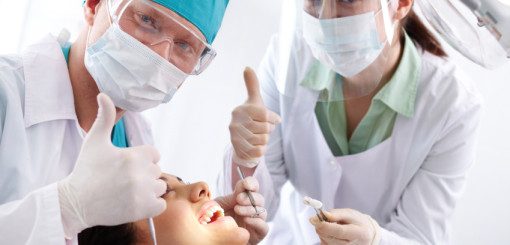 Two dental professionals, wearing masks and gloves, give thumbs-up while assisting a patient in a dental chair. The patient appears relaxed and is receiving treatment.