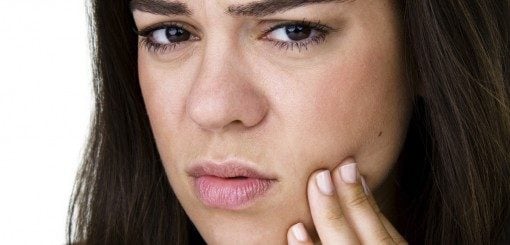 A young woman is touching her cheek with a concerned expression, indicating possible pain or discomfort. She has long, dark hair and is looking directly at the camera. The background is plain and light-colored.