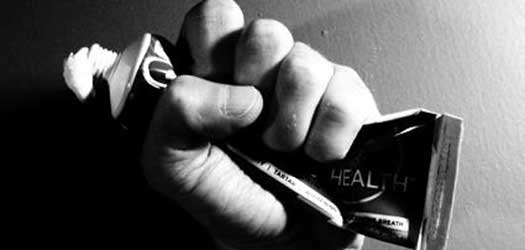 A hand grips a squeezed tube of toothpaste, with some paste emerging from the opening. The image is in black and white, emphasizing the tension and focus on the action of squeezing.