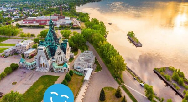 Vue aérienne d'un paysage urbain au bord d'une rivière avec une église historique, une végétation luxuriante et un cours d'eau réfléchissant sous un ciel vibrant.
