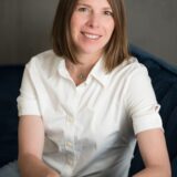 A smiling woman with shoulder-length brown hair is seated on a blue couch, wearing a white shirt and jeans, and has a casual, friendly demeanor.
