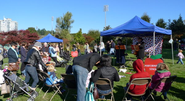 A crowd enjoys a lively outdoor event in a park, featuring a blue tent where musicians perform. Attendees, including families with children, are seated on chairs and lawn, surrounded by greenery and trees under a clear blue sky.