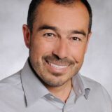 A smiling man with short dark hair and a neatly trimmed mustache, wearing a light gray shirt. The background is a soft gray, highlighting his friendly expression.