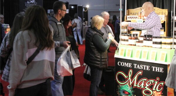 A lively market scene shows a vendor offering samples at a booth labeled "Come Taste the Magic." Customers engage with the vendor while exploring various products on display, surrounded by other stalls in a bustling event space with a vibrant red carpet.