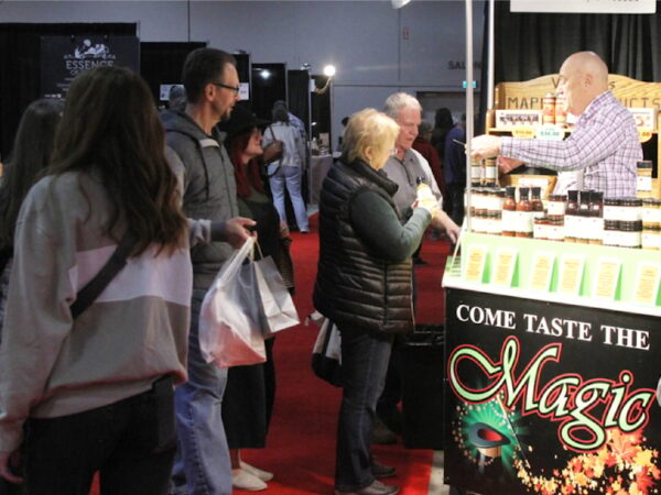 A lively market scene shows a vendor offering samples at a booth labeled "Come Taste the Magic." Customers engage with the vendor while exploring various products on display, surrounded by other stalls in a bustling event space with a vibrant red carpet.