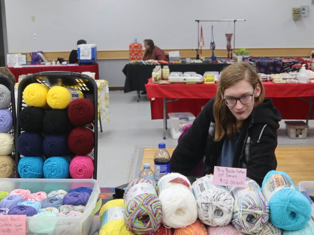 A person sits at a table filled with colorful yarn skeins, surrounded by additional vendor booths in a market setting. The atmosphere is vibrant and inviting, showcasing a variety of crafts and handmade goods.