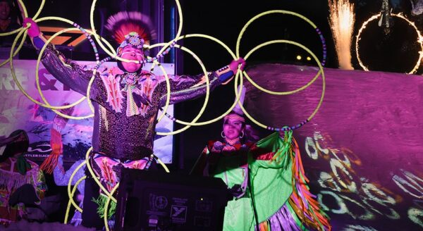 A performer dressed in colorful traditional attire showcases hoop dancing, surrounded by illuminated hoops, while another performer stands beside them on stage. The backdrop features vibrant lights and fireworks, creating a festive atmosphere.