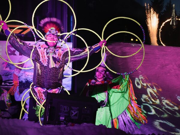 A performer dressed in colorful traditional attire showcases hoop dancing, surrounded by illuminated hoops, while another performer stands beside them on stage. The backdrop features vibrant lights and fireworks, creating a festive atmosphere.