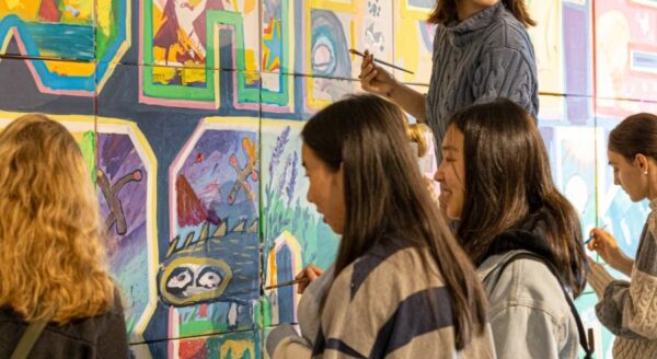 A group of young women is engaged in painting a colorful mural on a wall, with one woman smiling and holding a brush. The vibrant artwork features various designs and characters, showcasing creativity and collaboration among the participants.