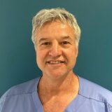 A smiling male doctor with gray hair, wearing a blue surgical scrub top, stands in front of a solid teal background.