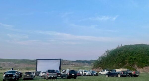 A group of parked vehicles faces a large outdoor movie screen set up in a grassy field, under a clear sky.