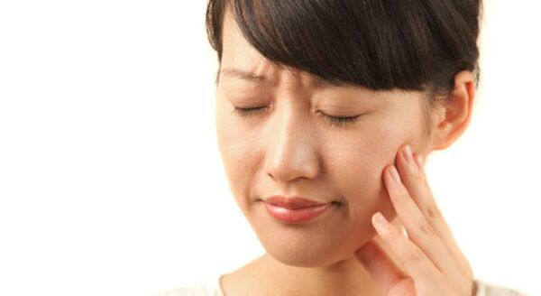 A woman with dark hair is shown holding her jaw with a pained expression, indicating discomfort. Her eyes are closed, and she appears to be experiencing dental or facial pain.