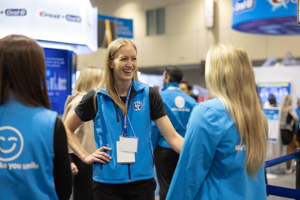A woman in a blue vest is engaged in conversation with two women at an event. The setting features a booth with informational displays. The friendly 123Dentist Smiley can be seen nearby, adding to the welcoming atmosphere.
