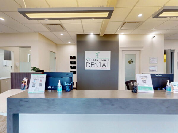 A dental office reception area featuring a modern design with a welcome desk displaying the sign "Village Mall Dental." The space is well-lit and includes informational brochures and hygiene products.